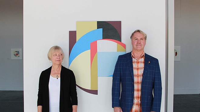 Artists Constance Lowe and Gary Sweeney stand stiffly in front of one of Lowe’s geometric abstractions.