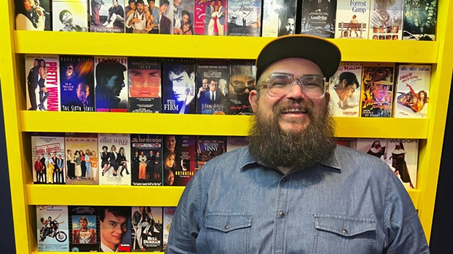Be Kind & Rewind owner Alex Amaro stands in front of a display of VHS covers inside the bar.